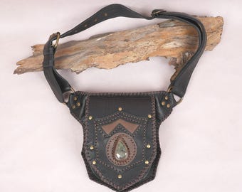 May include: A black leather hip bag featuring a shield-shaped front flap with brown leather accents, brass-toned metal rivets, and a central teardrop-shaped green stone inlay. The bag includes an adjustable leather strap with brass-toned hardware.