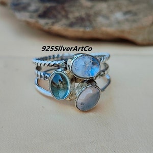 Puede incluir: Un anillo de plata con tres piedras preciosas ovaladas. El anillo está hecho de alambre de plata trenzado y presenta una piedra preciosa azul claro, una blanca y una rosa.