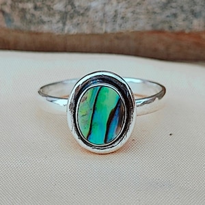 Puede incluir: Un anillo de plata con una incrustación ovalada de concha de abulón. La concha tiene un patrón iridiscente verde, azul y negro.