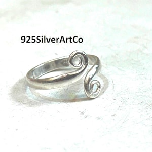 Può includere: Un anello d'argento con un design a spirale. L'anello presenta due decorazioni a spirale e il testo "925SilverArtCo". L'anello è posto su uno sfondo bianco.