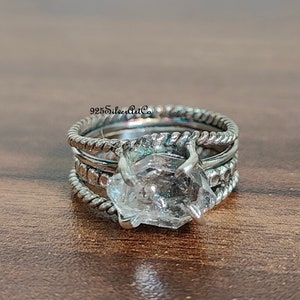 Peut inclure: Une bague en argent avec un grand cristal de quartz clair serti au centre. La bague est composée de plusieurs bandes, chacune avec une texture différente. La bague est posée sur une surface en bois.
