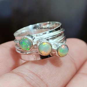 May include: A silver ring featuring three opal gemstones. The ring has a textured band, and the opals are round and set in individual bezels. The opals display a play of colour with flashes of green, yellow, and orange. The text "925SilverArtCo" is visible.