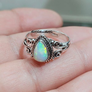 May include: A silver ring with a pear-shaped opal gemstone. The ring has a delicate, ornate design with swirls and a rope-like band. The opal is iridescent with shades of white, green, and blue.