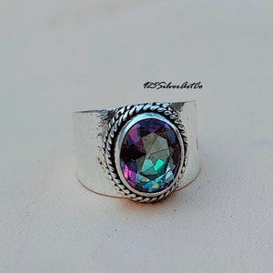 May include: A wide silver ring featuring an oval-cut, iridescent gemstone. The gemstone displays a spectrum of colors, including purple, green, and blue. The ring has a textured, rope-like border around the stone and a wide band. The text "425silverartco" is visible.