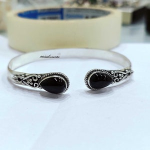 May include: A silver bangle bracelet with two black teardrop-shaped stones. The bracelet has a delicate, ornate design.