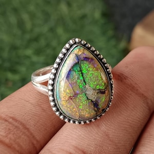 May include: A teardrop-shaped ring featuring an iridescent gemstone. The stone has green, gold, and purple colours, set in a silver band with a beaded edge. A unique piece of jewellery.