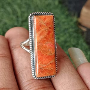 May include: A rectangular orange gemstone ring with a silver band. The gemstone has a textured surface and is set within a silver frame with a rope-like border. The ring is displayed on a hand.