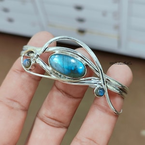 Op de afbeelding: Een zilveren armband met een blauwe labradorit edelsteen en twee kleinere ronde edelstenen. De armband heeft een delicaat, ingewikkeld ontwerp.