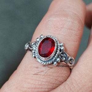 May include: A silver ring with a red gemstone set in a detailed oval bezel. The ring has a delicate, ornate design with swirls and small beads.