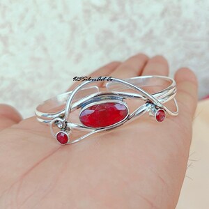 May include: Silver bangle bracelet with a red gemstone and two smaller red gemstones. The bracelet is open and adjustable.