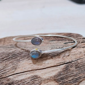 May include: A silver bangle bracelet with two round labradorite gemstones. The bracelet is open and has a simple design.
