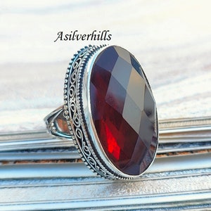 May include: A silver ring featuring an intricate design and a large, faceted, dark red gemstone.