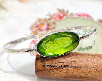 May include: A silver bangle bracelet featuring a large, oval-shaped green gemstone. The bracelet is displayed on a wooden surface.