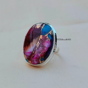 May include: A silver ring with a large oval-shaped gemstone. The gemstone is a mix of pink, purple, brown, and blue colors with gold flecks.