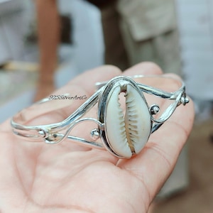 May include: A silver-toned cuff bracelet featuring a natural cowrie shell centerpiece. The shell is cream-colored with a textured surface, set within an oval frame. The bracelet has a delicate, open design with decorative swirls and the text "925SilverArtCo".