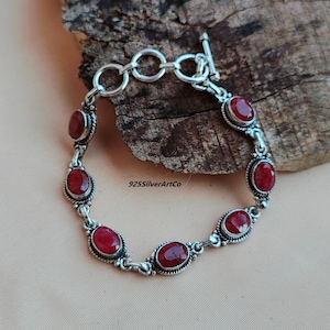 May include: A silver bracelet with a toggle clasp and six red oval gemstones. The bracelet is on a wooden surface.