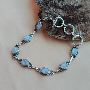May include: A silver bracelet with a toggle clasp and five oval moonstone gemstones. The bracelet is on a light brown wooden surface.
