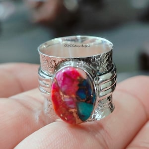 May include: A silver ring with a colorful, oval-shaped gemstone set in the center. The band is textured and has a hammered finish. The ring is inscribed with "925SilverArtCo."