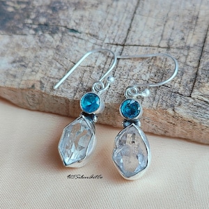 May include: Silver dangle earrings with a clear quartz crystal and a blue gemstone. The earrings have a simple design and are perfect for everyday wear.