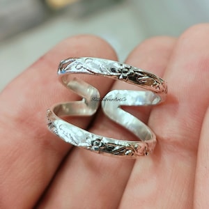 May include: A silver ring with a floral design. The ring is made of two bands that are intertwined. The ring is worn on a finger.