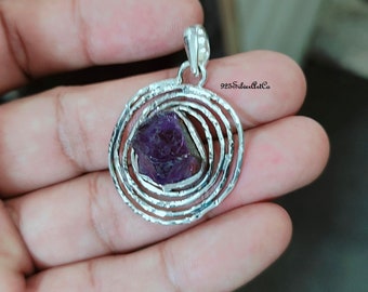 May include: A silver pendant with a spiral design and a purple amethyst stone in the center. 925SilverArtCo is printed on the pendant.