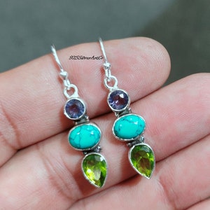 May include: Silver dangle earrings with three gemstones. The top stone is a small round purple amethyst, the middle stone is a turquoise oval, and the bottom stone is a pear-shaped green peridot.