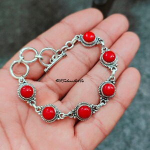 May include: A silver bracelet with a toggle clasp and six red coral stones. The bracelet is on a hand.