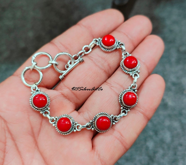 May include: A silver bracelet with a toggle clasp and six red coral stones. The bracelet is on a hand.