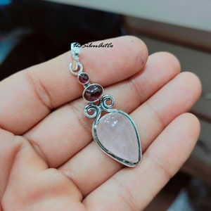 May include: A silver pendant with a pink rose quartz stone and two smaller garnet stones. The pendant has a delicate, ornate design.