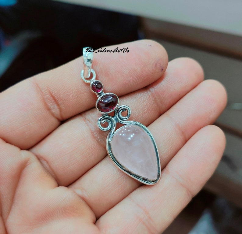 May include: A silver pendant with a pink rose quartz stone and two smaller garnet stones. The pendant has a delicate, ornate design.