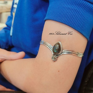 May include: A silver arm cuff with a large teardrop-shaped gemstone and a smaller round gemstone. The cuff is on a person's arm and the text "925 Silverart Co." is written on the skin.