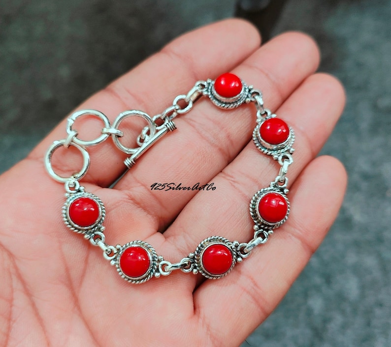May include: A silver bracelet with a toggle clasp and six red coral stones. The bracelet is on a hand.