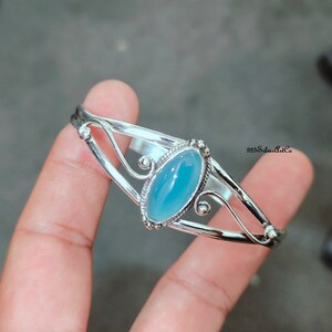 K&ouml;nnte beinhalten: Ein silbernes Armreif-Armband mit einem hellblauen ovalen Edelstein, der in einem dekorativen silbernen Rahmen gefasst ist. Das Armband hat ein zartes, filigranes Design.