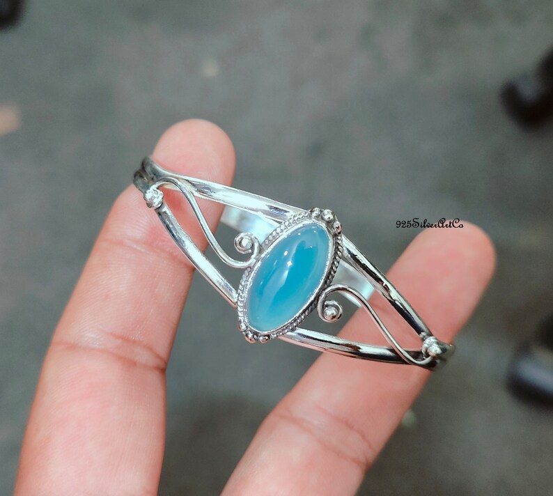 K&ouml;nnte beinhalten: Ein silbernes Armreif-Armband mit einem hellblauen ovalen Edelstein, der in einem dekorativen silbernen Rahmen gefasst ist. Das Armband hat ein zartes, filigranes Design.