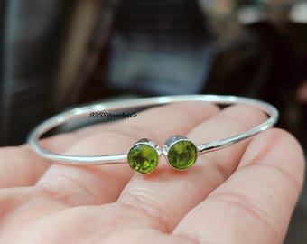 May include: A silver bangle bracelet with two green gemstone accents. The bracelet is open and has a simple design.
