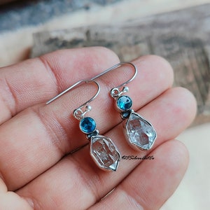 May include: Silver dangle earrings with a clear quartz teardrop and a small blue gemstone. The earrings have a hook closure.