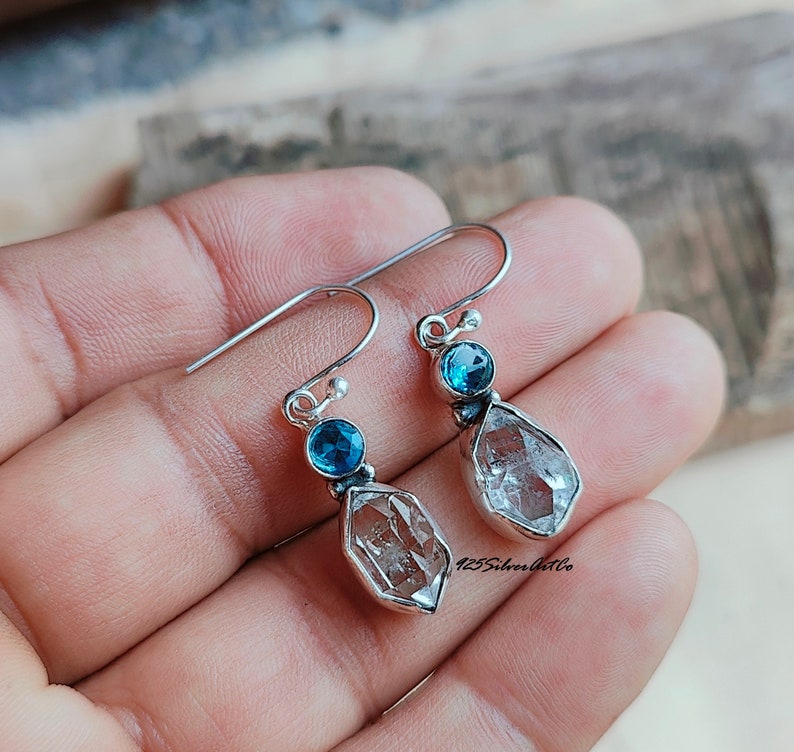 May include: Silver dangle earrings with a clear quartz teardrop and a small blue gemstone. The earrings have a hook closure.