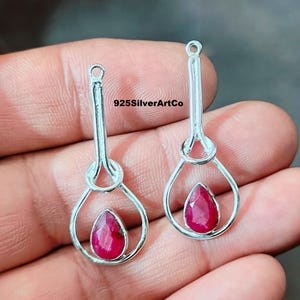 May include: A pair of silver earrings with a teardrop-shaped ruby gemstone in each. The earrings have a delicate, knotted design and are perfect for adding a touch of elegance to any outfit.