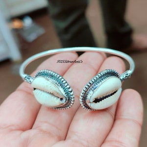 May include: Silver bangle bracelet with two white seashells. The bracelet is decorated with a beaded border around each seashell. The text "925SilverArtCo" is visible on the bracelet.