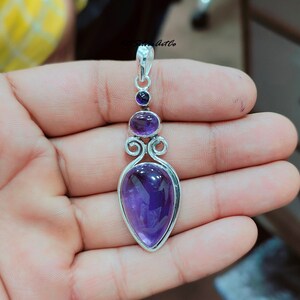 May include: A silver pendant with a teardrop-shaped amethyst gemstone. The pendant has a delicate design with two smaller amethyst gemstones above the main stone.