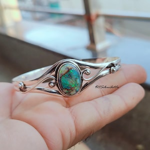May include: A silver bracelet with a green and white opal gemstone set in a silver oval. The bracelet has a delicate, swirling design.