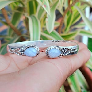 Könnte beinhalten: Ein silbernes Armreif-Armband mit zwei tropfenförmigen blauen Achat-Steinen. Das Armband hat ein zartes Design mit kleinen silbernen Perlen und Schnörkeln.