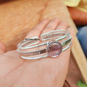 May include: Silver bangle bracelet with a round, light purple gemstone. The bracelet has a textured, ribbed design.
