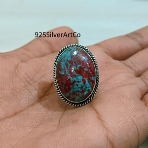 May include: A silver ring with a red and blue oval-shaped gemstone set in a silver bezel. The ring is on a hand.