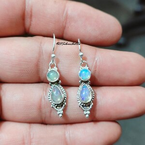 May include: A pair of silver earrings with opal gemstones. Each earring features a small round opal at the top and a larger, teardrop-shaped opal below. The earrings have a decorative, textured border and a hook closure.