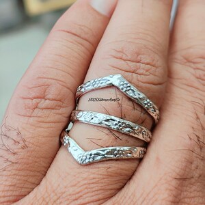 Op de afbeelding: Een zilveren ring met een chevron-ontwerp. De ring is gemaakt van drie banden die op elkaar zijn gestapeld. De banden zijn versierd met een bloemmotief. De ring wordt aan een vinger gedragen.