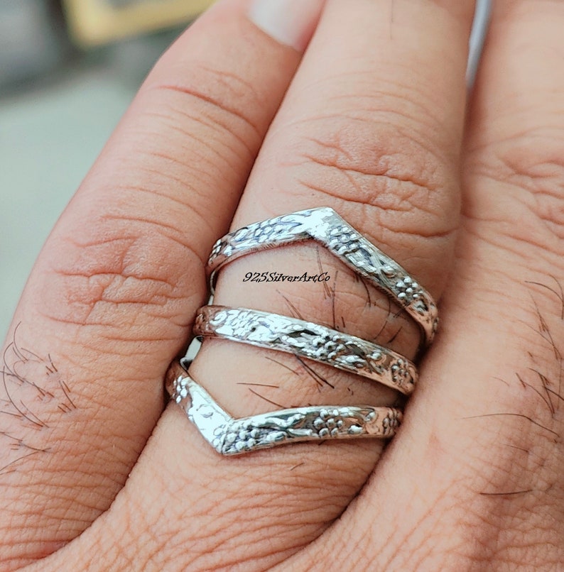 Op de afbeelding: Een zilveren ring met een chevron-ontwerp. De ring is gemaakt van drie banden die op elkaar zijn gestapeld. De banden zijn versierd met een bloemmotief. De ring wordt aan een vinger gedragen.