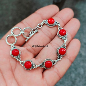 May include: A silver bracelet with a toggle clasp and six red coral stones. The bracelet is on a hand.