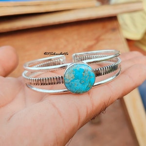 May include: A silver cuff bracelet with a turquoise stone set in the center. The bracelet has a textured design and three bands.