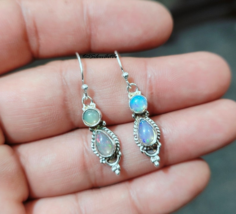May include: A pair of silver earrings with a teardrop design. Each earring features a pearlescent opal stone and a smaller, round, light green stone. The earrings are displayed against a neutral background.